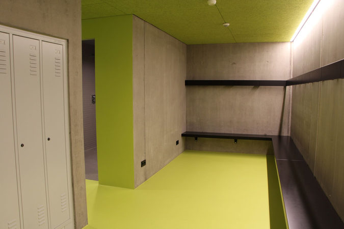 Ein Umkleideraum mit grauen Wänden, einem hellgrünen Boden und Decke, schwarzen Bänken an den Wänden und weißen Schließfächern links.