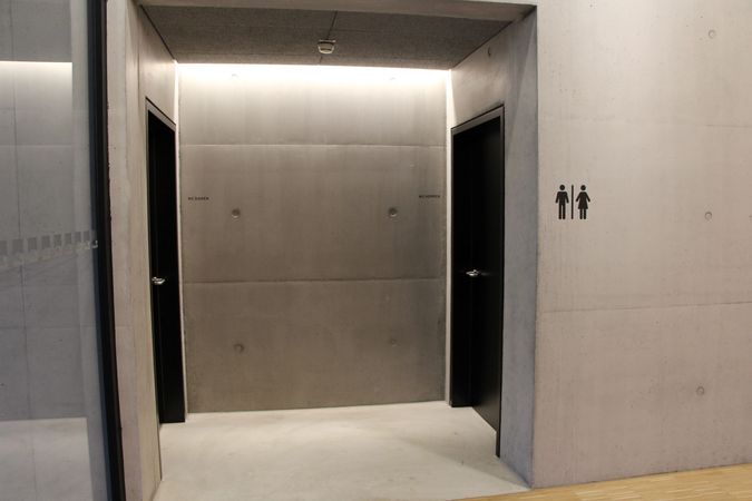 Ein grauer, minimalistischer Flur mit zwei Türen zu den Toiletten, gekennzeichnet durch Piktogramme für Männer und Frauen an der Wand.