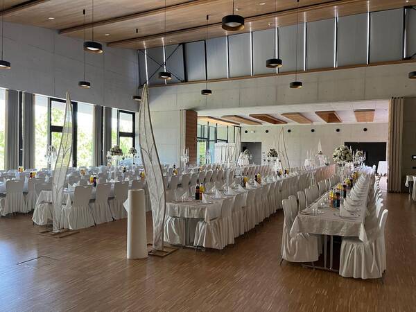 Ein großer Festsaal ist elegant für ein Event mit langen, weiß gedeckten Tischen und Stühlen vorbereitet.