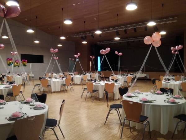 Ein festlich dekorierter Saal mit runden, weiß gedeckten Tischen und rosa Luftballons, die von der Decke hängen.