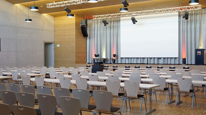 Ein großer, leerer Konferenzraum mit Stuhlreihen und Tischen ist auf eine Bühne mit einem weißen Projektionsbildschirm ausgerichtet.