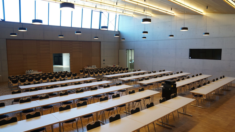 Ein großer, moderner Konferenzraum mit vielen länglichen Tischen und Stühlen ist leer und von einer hohen Decke mit hängenden Lampen beleuchtet.