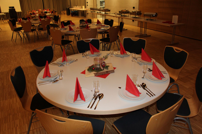 Ein großer, runder Tisch in einem festlich dekorierten Raum ist für ein Essen gedeckt, mit roten Servietten, glänzendem Besteck und einem weihnachtlichen Mittelstück.