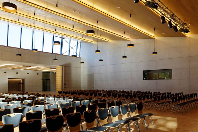 Ein moderner, geräumiger Saal mit vielen Stuhlreihen, einer hohen Decke und großen Fenstern, die den Raum erhellen.