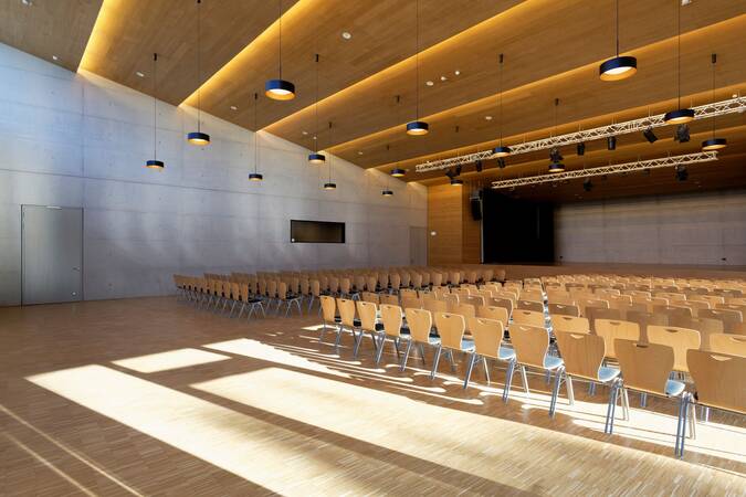 Ein moderner, leerer Veranstaltungssaal mit Holzboden, hölzerner Decke und mehreren Reihen aneinander gereihter Holzstühle ist von warmem, einfallendem Licht erleuchtet.