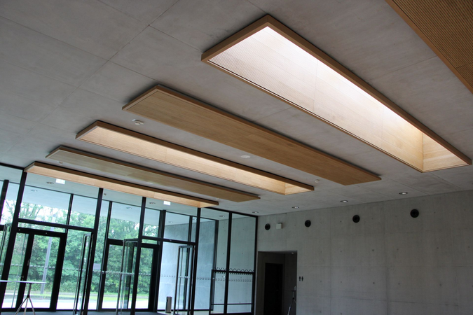Die Decke eines modernen Innenraums wird von mehreren rechteckigen Lichtfeldern in Holzoptik beleuchtet, während große Fenster im Hintergrund den Blick nach draußen freigeben.