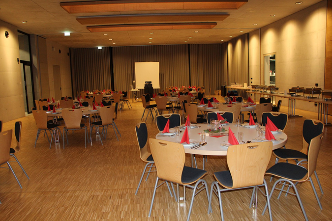In einem großen Saal stehen mehrere runde Tische mit Holzstühlen und Gedecken, auf denen rote Servietten platziert sind, und im Hintergrund befinden sich ein Buffetbereich sowie ein Projektionsschirm.
