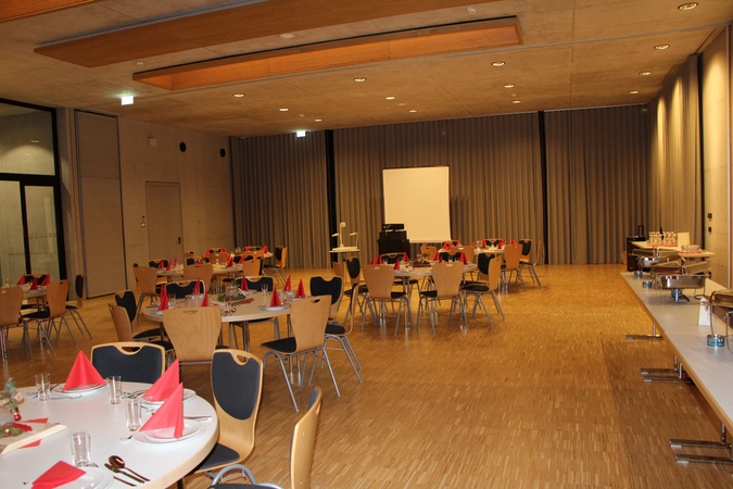 Ein großer, festlich eingedeckter Saal mit runden Tischen, roten Servietten und einer Leinwand im Hintergrund.