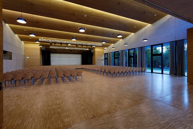 In einem modernen, geräumigen Saal mit Holzverkleidung stehen viele leere Stuhlreihen vor einer Bühne, während große Glasfenster auf der rechten Seite den Blick in einen Außenbereich ermöglichen.