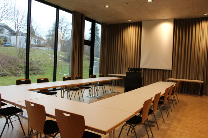 Ein Konferenzraum mit U-förmig angeordneten Tischen und leeren Stühlen, großen Fenstern mit Blick nach draußen und einem weißen Projektionsschirm an der Wand.