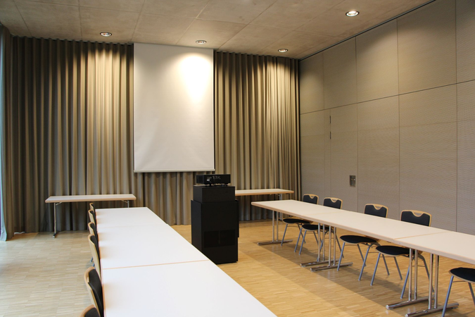 Der Konferenzraum ist mit Tischen und Stühlen in U-Form eingerichtet, hat Vorhänge an den Fenstern und eine Leinwand an der Wand.