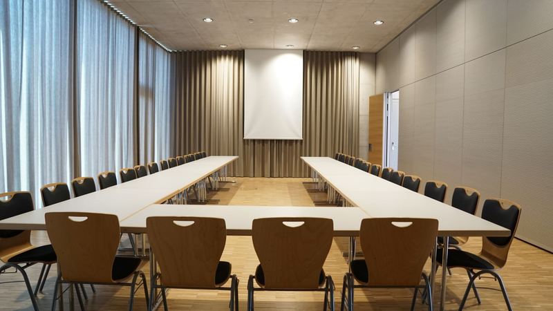 Ein leerer Konferenzraum mit einem U-förmig angeordneten Tisch, umgeben von Stühlen, vor einem geschlossenen Vorhang und einer Leinwand.