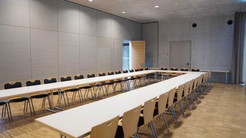 Der Raum zeigt einen großen, leeren Konferenzsaal mit Tischen in U-Form und vielen Stühlen auf hellem Holzboden.