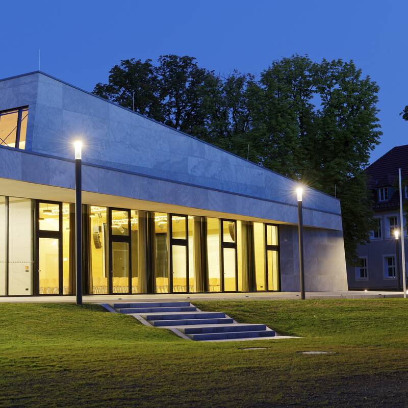 Ein modernes, beleuchtetes Gebäude mit großen Glasfenstern steht neben grünen Bäumen im Abendlicht.
