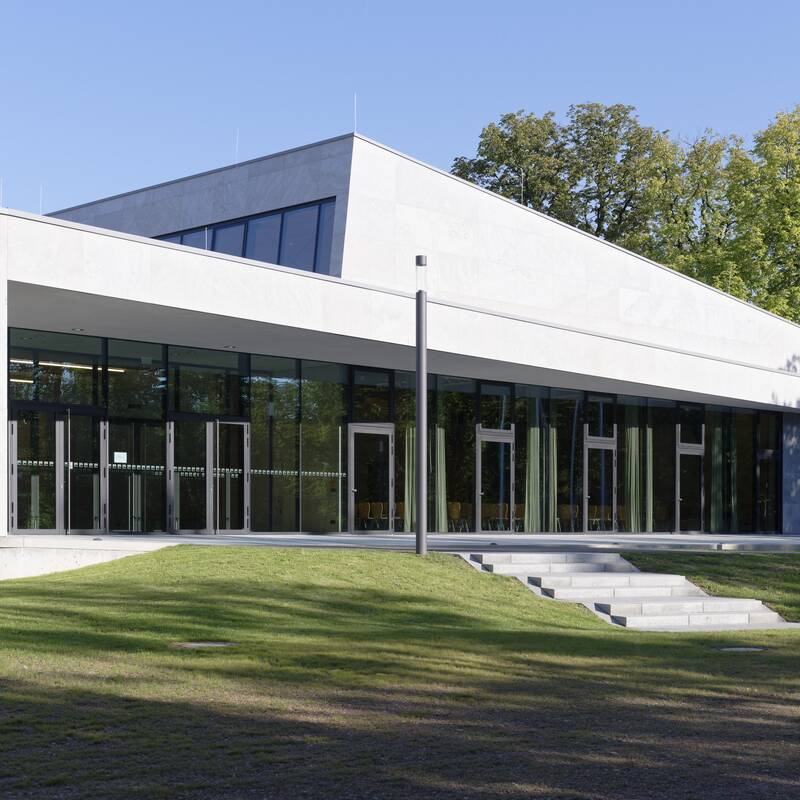 Ein modernes Gebäude mit großen Glasfenstern steht vor einer Grünfläche, umgeben von Bäumen.